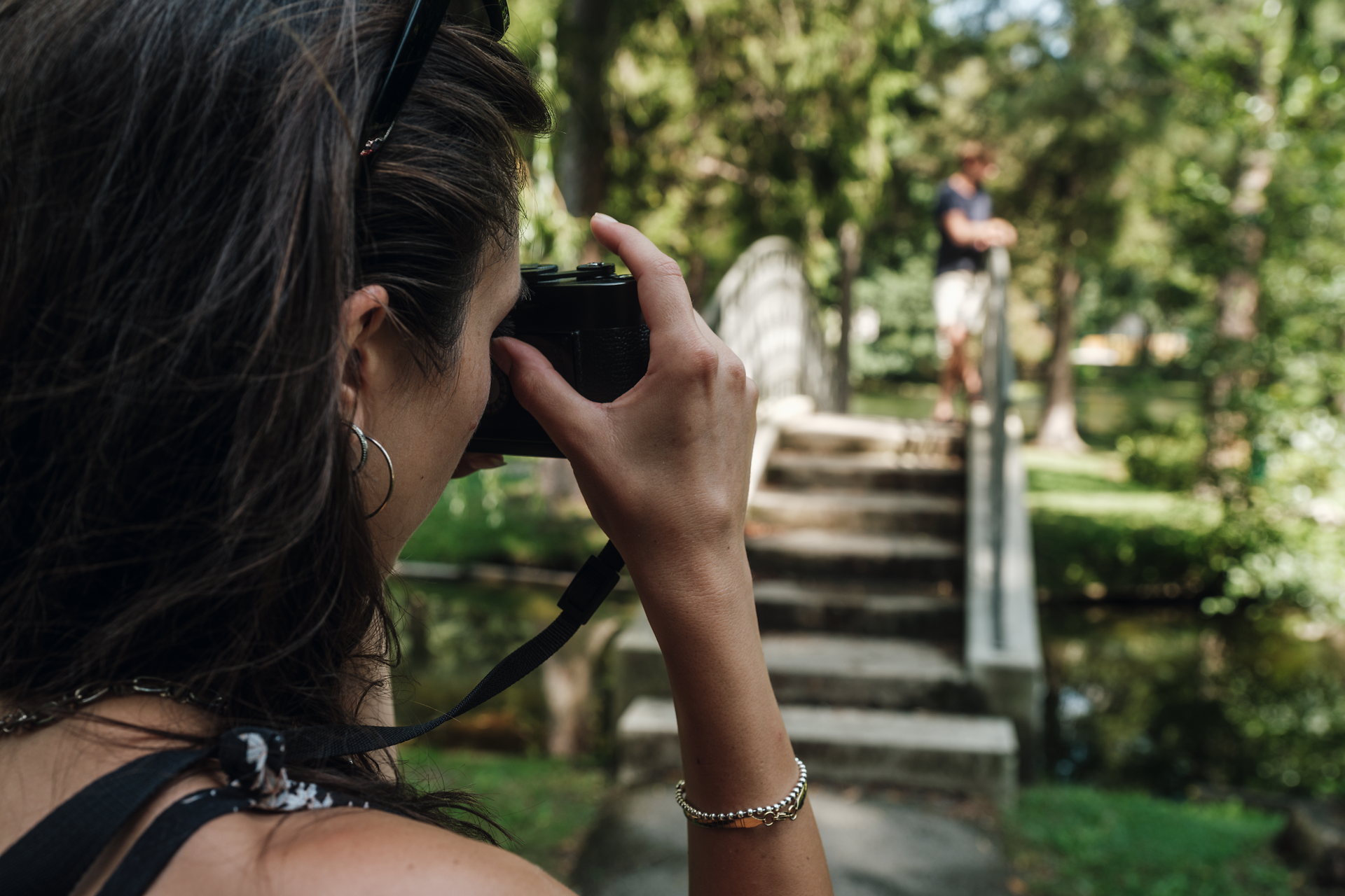 Inmitten einer malerischen Landschaft hält eine Fotografin den Moment fest, während ihr Freund auf einer charmanten Brücke posiert. Umgeben von üppigem Grün und dem sanften Plätschern des Wassers, strahlt die Szene eine entspannte Sommeratmosphäre aus, die zum Verweilen einlädt.