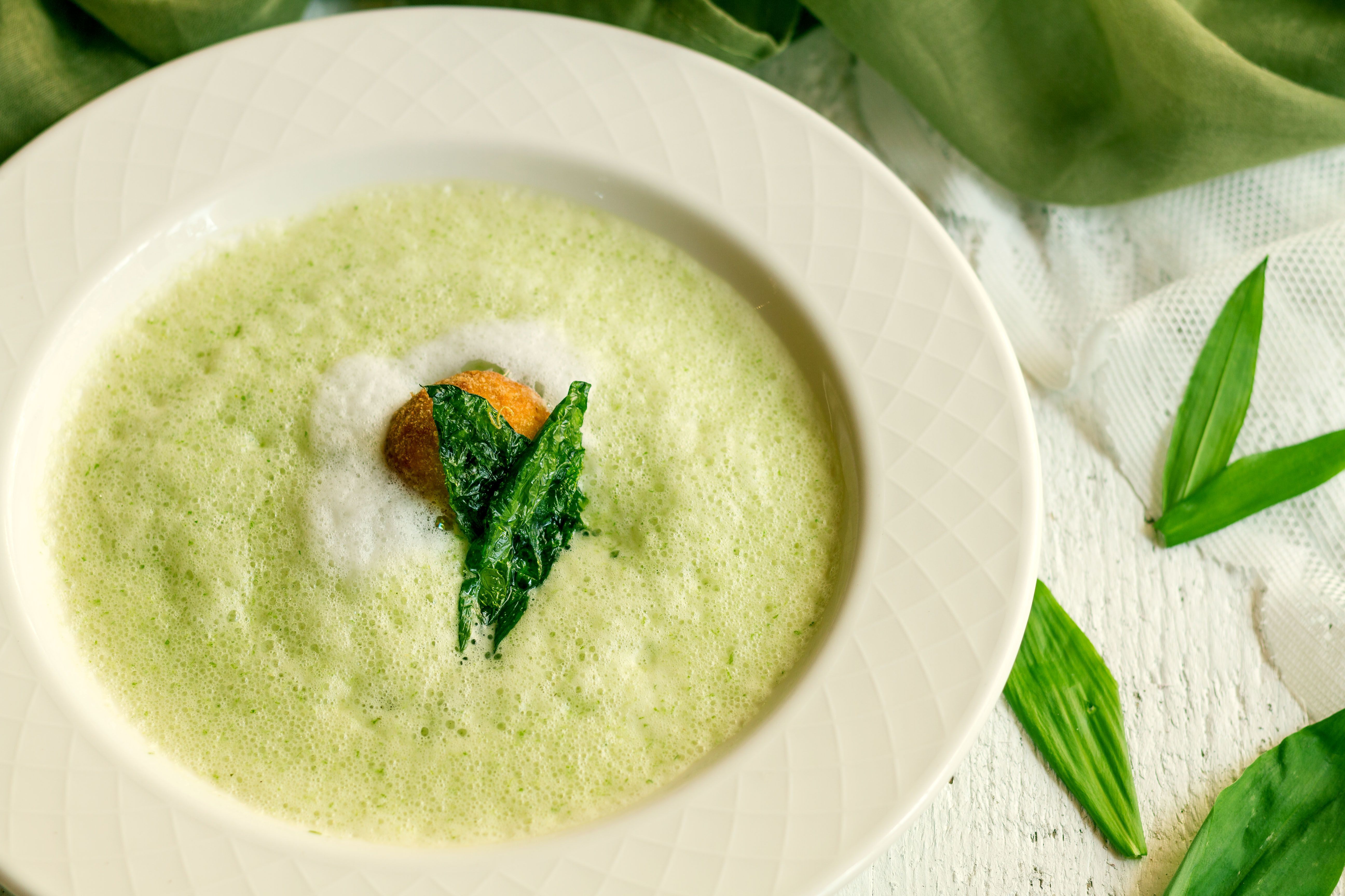 Ein weißes Teller mit Berlauchschaumsuppe, daneben Bärlauchblätter.