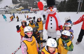 Kinder in Skiausrüstung posieren mit einem Schneemann auf einer Skipiste.