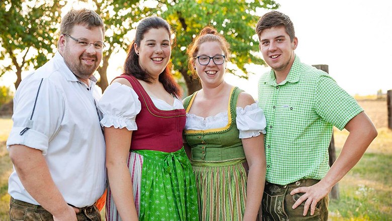 Four people in traditional costume stand smiling outdoors on a green meadow, with a tree in the background.