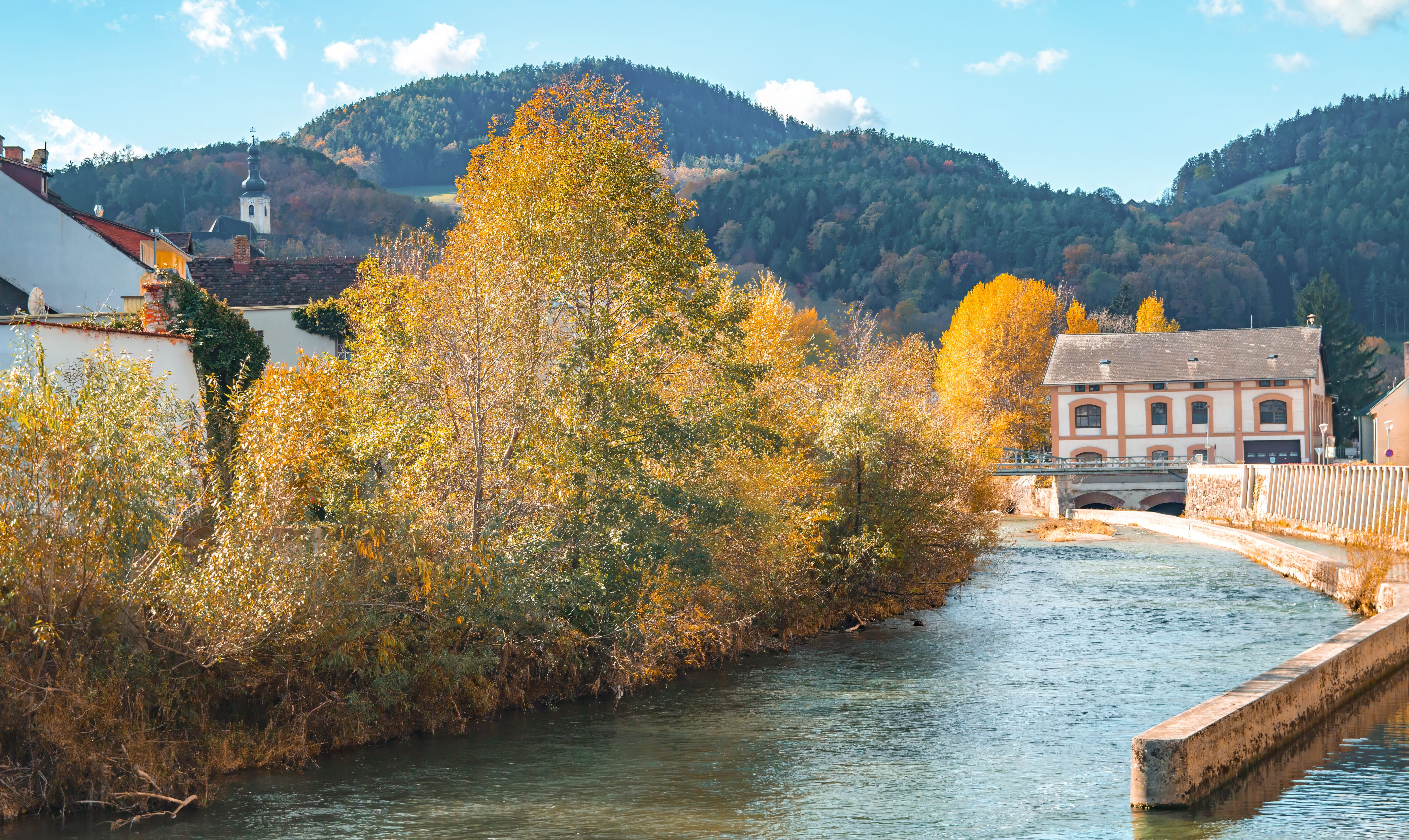 Fluss mit Herbstbäumen und Gebäude im Hintergrund, umgeben von Hügeln.