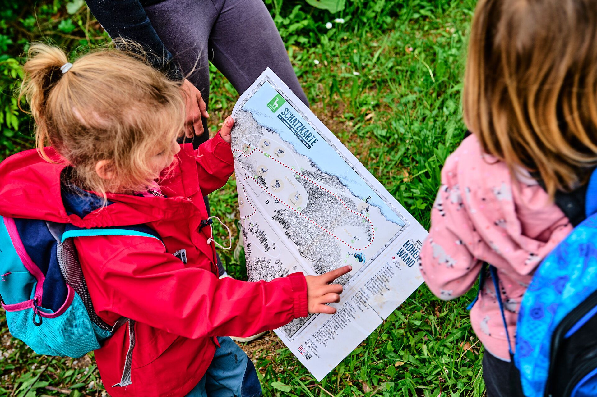 Zwei Kinder erkunden neugierig eine Wanderkarte, während die sanften Hügel und die üppige Natur um sie herum eine einladende Atmosphäre schaffen. Die strahlenden Farben ihrer Jacken heben sich lebhaft von der grünen Wiese ab, während sie sich auf ein Abenteuer in der Natur vorbereiten.