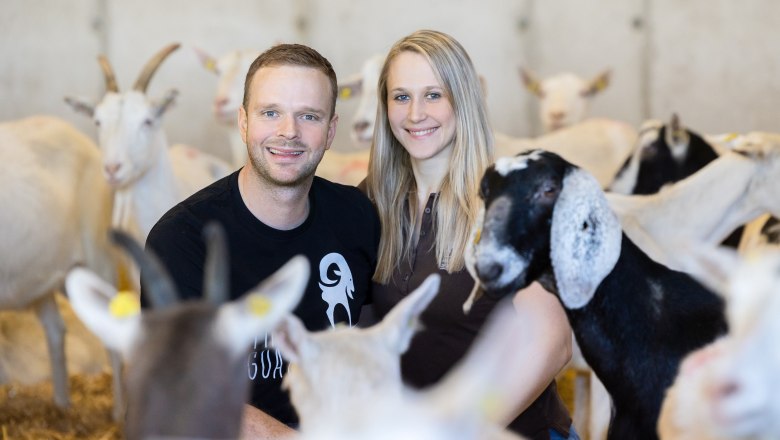 A man and a woman smile amidst goats in a stable.