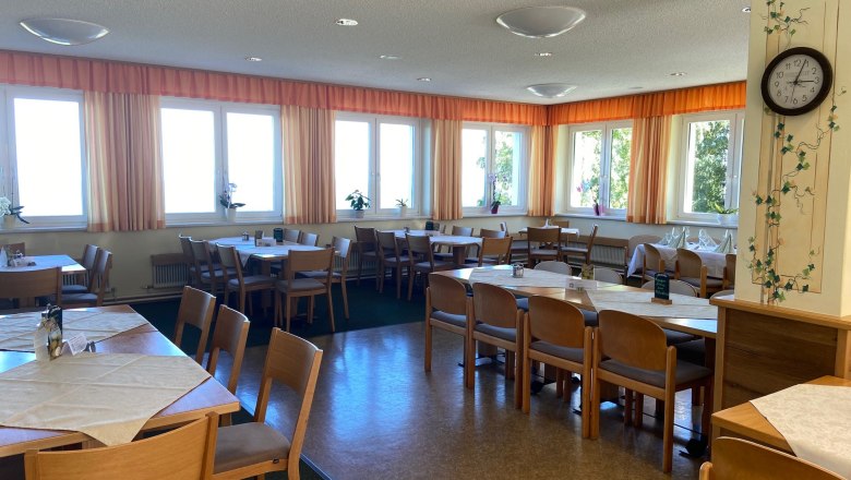 Ein heller Speisesaal mit Holzm&ouml;beln, Tischen und St&uuml;hlen, dekoriert mit Pflanzen und einer Wanduhr.