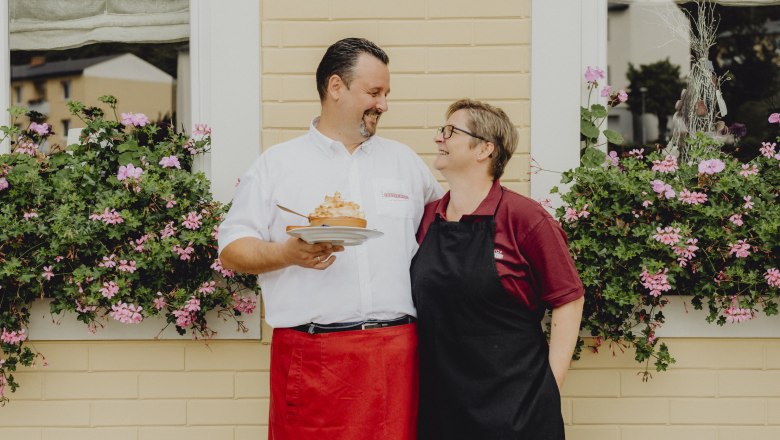 A man and a woman stand smiling in front of a window with flowers.