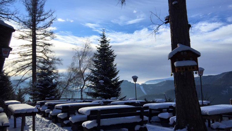 Terrasse Speckbacher Hütte, © Karin Stranz Verschneite Terrasse mit Holzbänken und Tischen, umgeben von Bäumen und Bergen im Hintergrund.