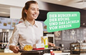 Eine l&auml;chelnde Frau in einem Fast-Food-Restaurant tr&auml;gt ein Tablett mit Pommes, einem Getr&auml;nk und einer Burger-Box. Text: 'Der Burger aus der K&uuml;che. Und das L&auml;cheln von Herzen.'