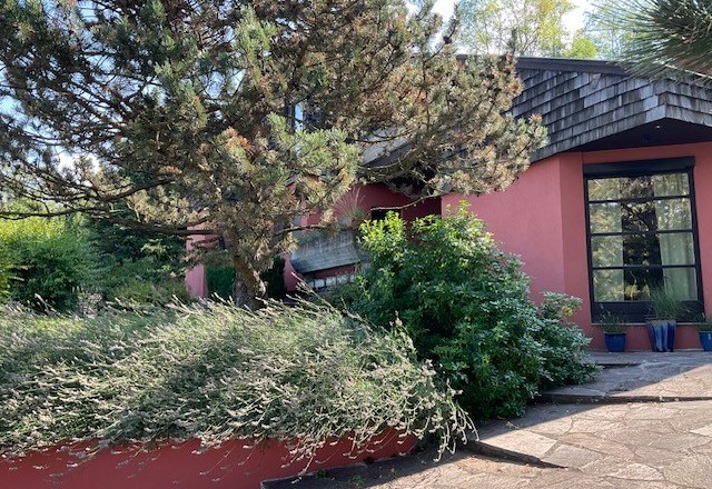 House view, © Renate Schrottenbach A modern house with a red façade, surrounded by trees and shrubs.