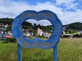 Blick durch einen blauen Rahmen mit der Aufschrift 'Sonnige Gr&uuml;&szlig;e aus Hollenthon' auf eine malerische Landschaft mit Dorf und Kirche.