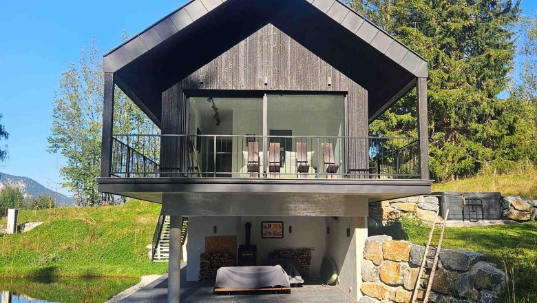 Modernes Haus mit Holzfassade und Balkon, umgeben von Natur und einem kleinen Teich.