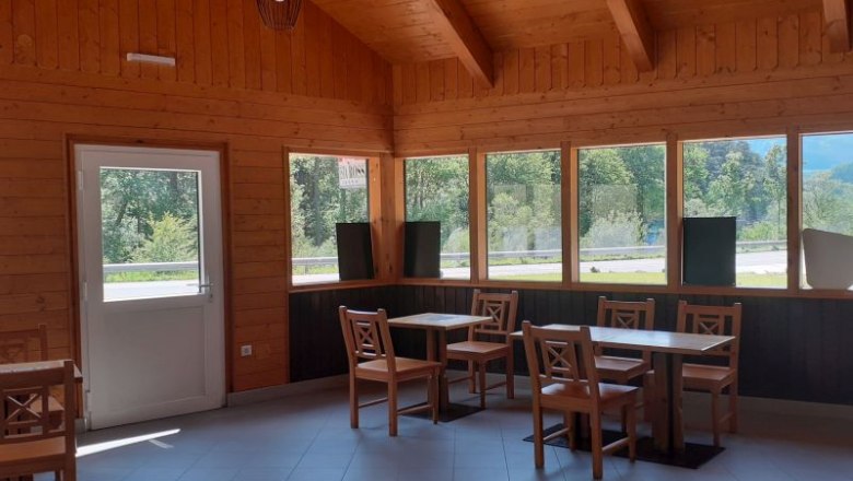 Innenansicht eines Holzgebäudes mit Tischen und Stühlen, großen Fenstern und einer Tür.