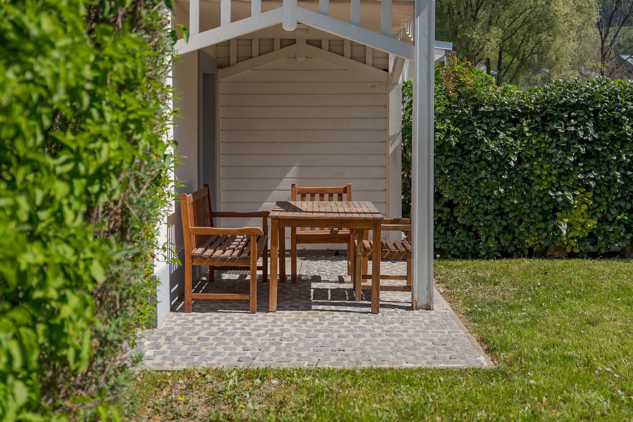 Ein Holztisch mit Stühlen steht unter einem kleinen Pavillon im Garten, umgeben von grünen Hecken und Rasen.