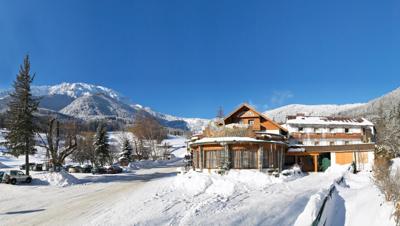 Winter landscape with Hotel Forellenhof in front of snow-covered mountains and a blue sky.