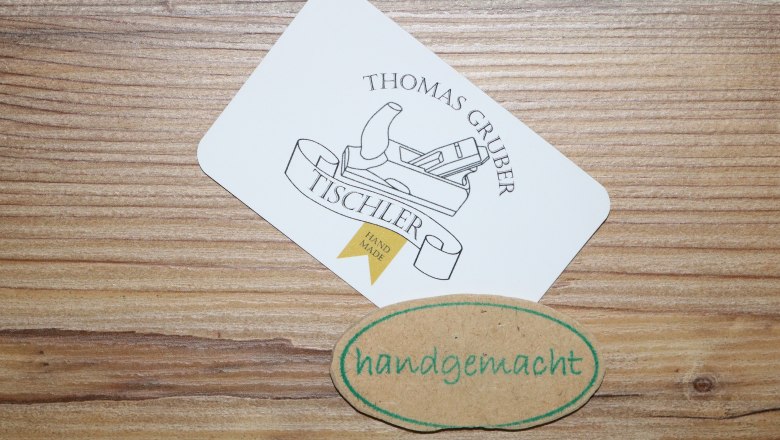 Visitenkarte von Tischler Thomas Gruber mit einem Hobel-Logo und einem handgemachten Etikett auf Holzuntergrund.