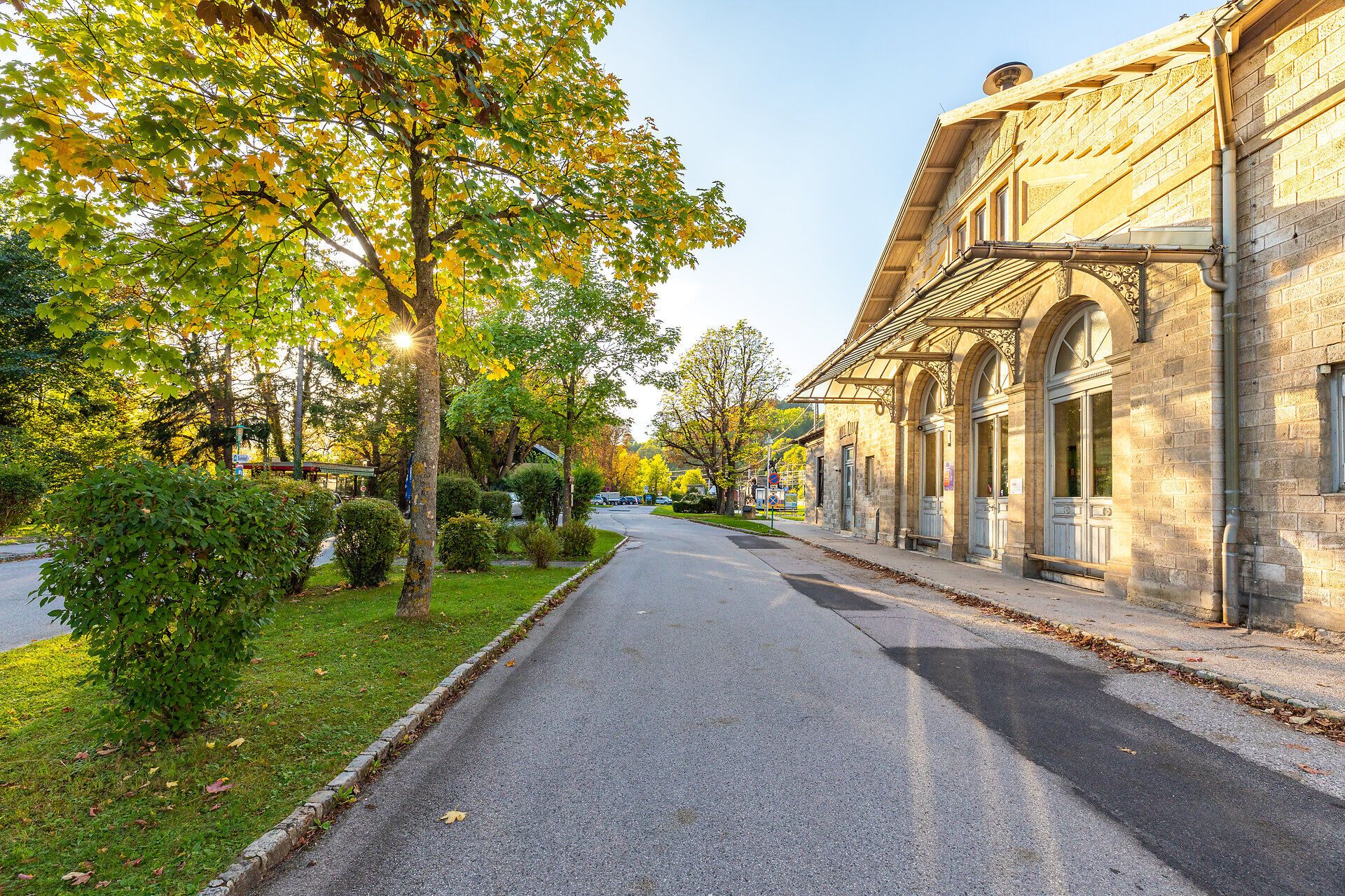 Das Bahnhofsgebäude von Payerbach wird von der Sonne angestrahlt, links sieht man eine Grünfläche mit Bäumen und Sträuchern.
