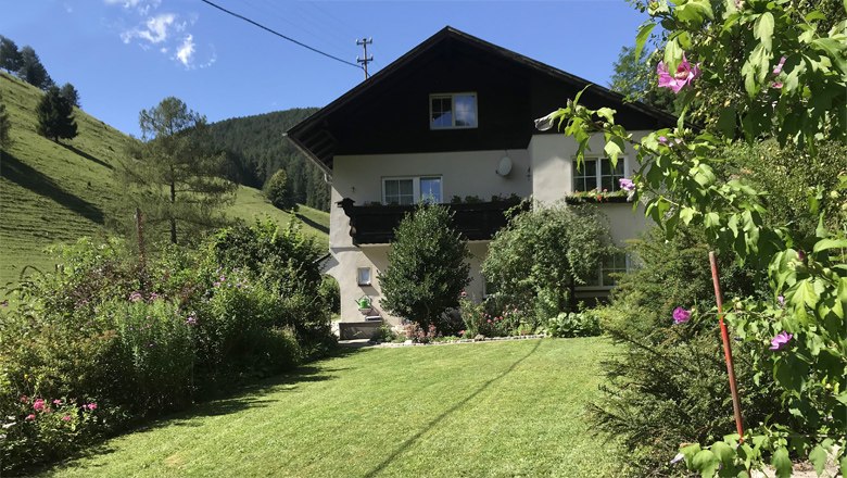 Ein zweistöckiges Haus mit Balkon, umgeben von einem gepflegten Garten und grünen Hügeln im Hintergrund.