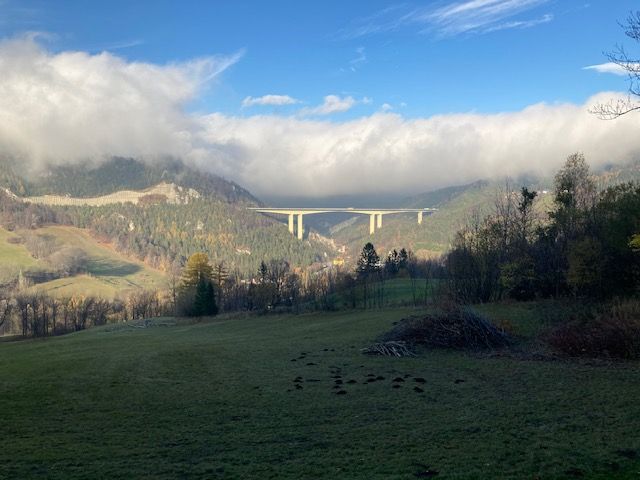 Landschaft mit Brücke über ein Tal, umgeben von bewaldeten Hügeln und Wolken.