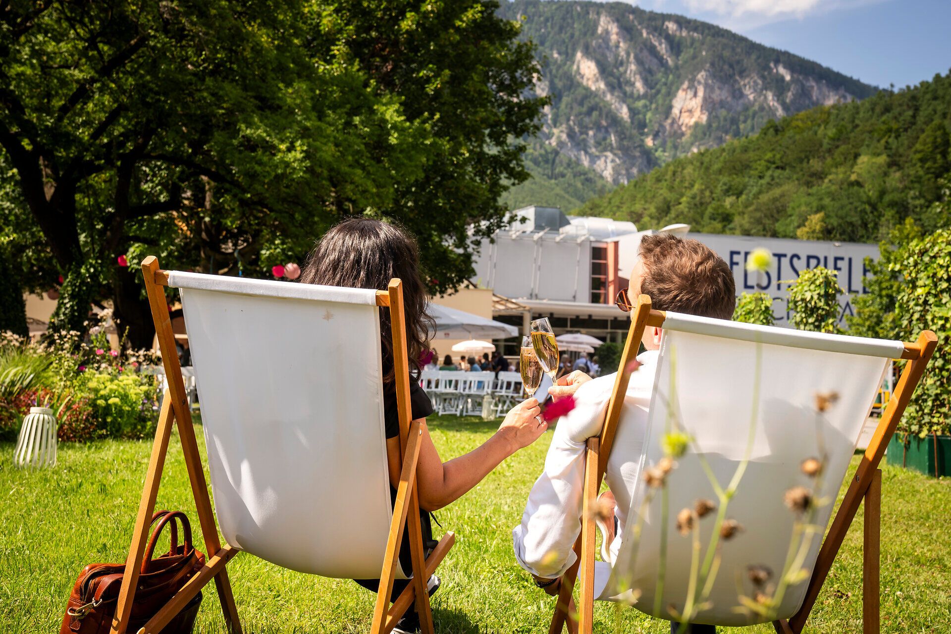 Inmitten der malerischen Kulisse der Wiener Alpen genießen zwei Personen in bequemen Liegestühlen ein Glas Sekt. Die sanften Hügel und die üppige grüne Wiese schaffen eine entspannte Atmosphäre, die zum Verweilen einlädt.