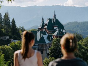 Zwei Menschen, mit dem Rücken zur Kamera, blicken auf eine malerische Berglandschaft. Im Vordergrund sieht man das Südbahnhotel, eingebettet in üppiges Grün. Im Hintergrund erheben sich Berge unter einem klaren blauen Himmel.