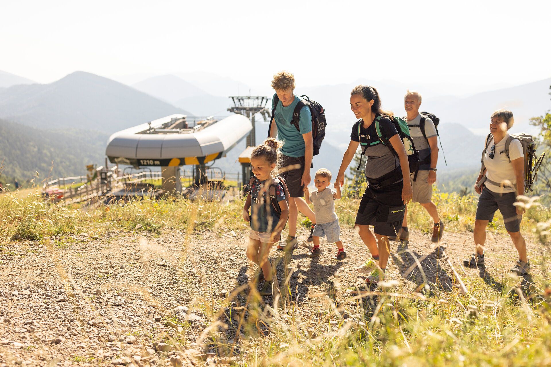 Eine fröhliche Wandergruppe erkundet die malerische Landschaft, während die sanften Hügel der Wiener Alpen im Hintergrund aufragen. Die Kinder lachen und spielen, während die Erwachsenen die frische Bergluft genießen und die Schönheit der Natur bewundern.