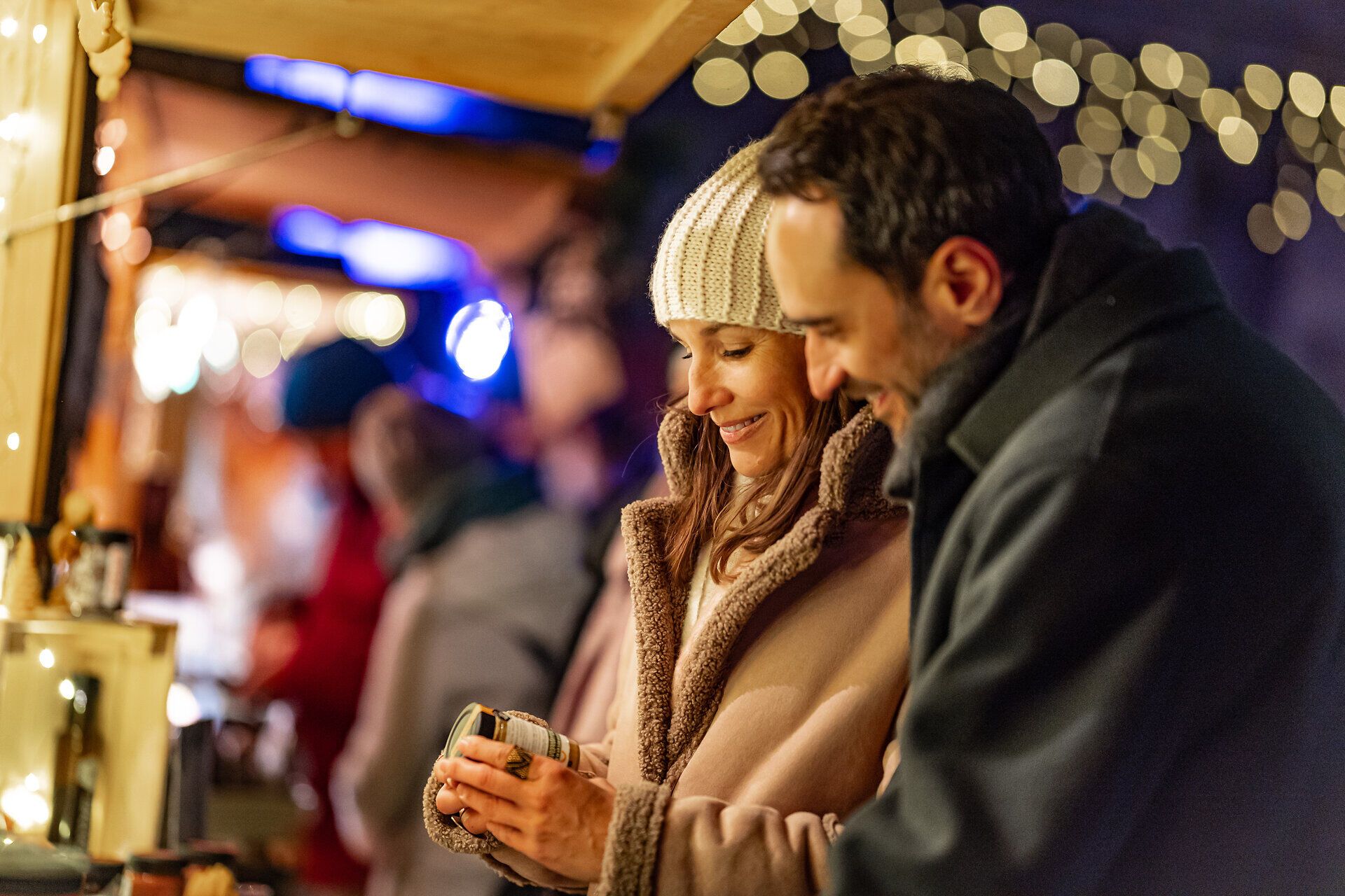 Paar betrachtet lächelnd eine Glas an einem beleuchteten Marktstand bei Nacht.