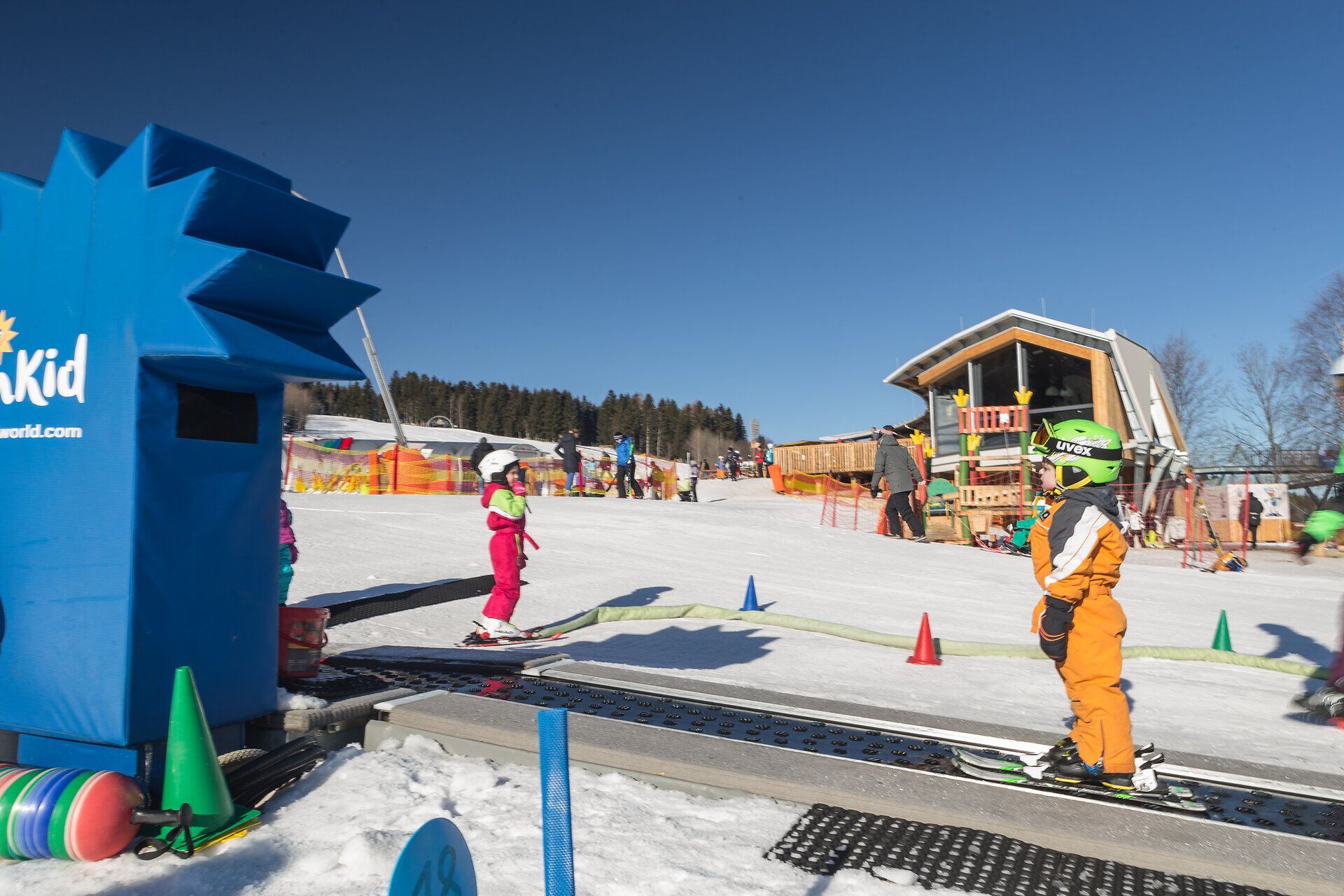 Ein strahlend blauer Himmel über einem lebhaften Skigebiet, wo Kinder fröhlich ihre ersten Schwünge auf den Pisten ziehen. Die bunten Skianzüge der kleinen Abenteurer bringen Freude und Energie in die winterliche Landschaft, während die frische Bergluft zum Lachen und Spielen einlädt.