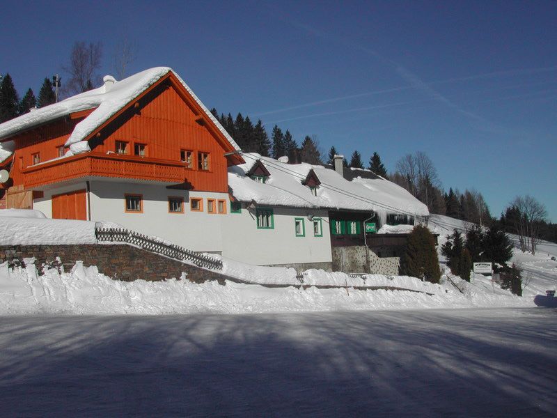 Ein verschneites Gasthaus mit rotem Holzgiebel und weißen Wänden am Feistritzsattel, umgeben von Bäumen und blauem Himmel.