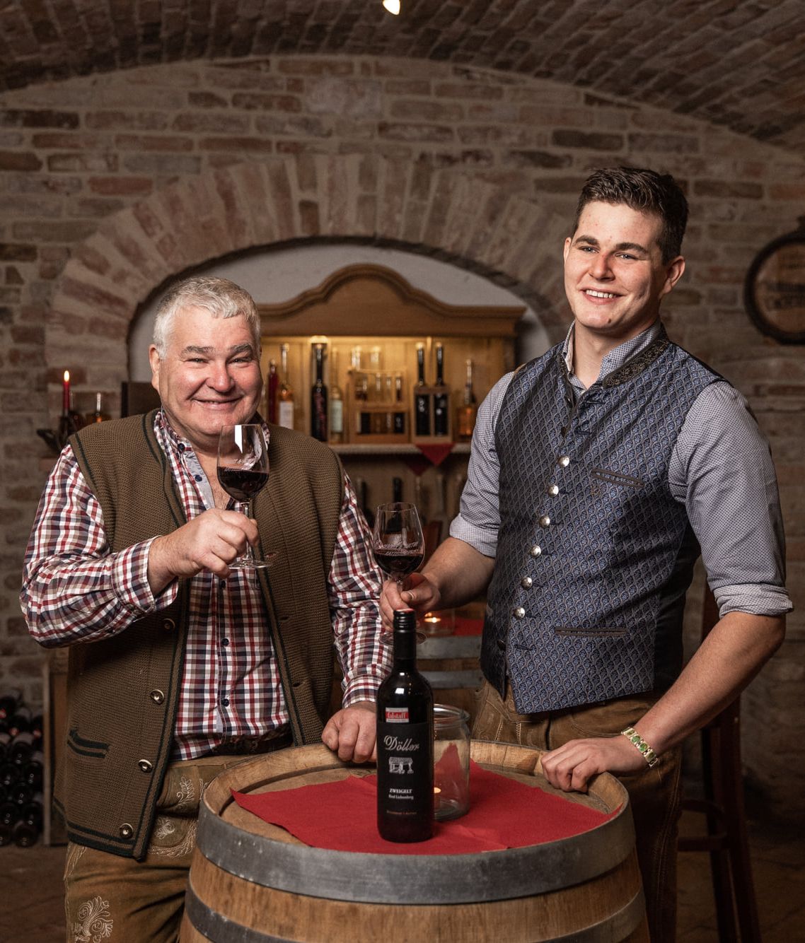 Ein junger und ein älterer Mann in einem Weinkeller mit Weingläsern in der Hand vor einem Holzfass.
