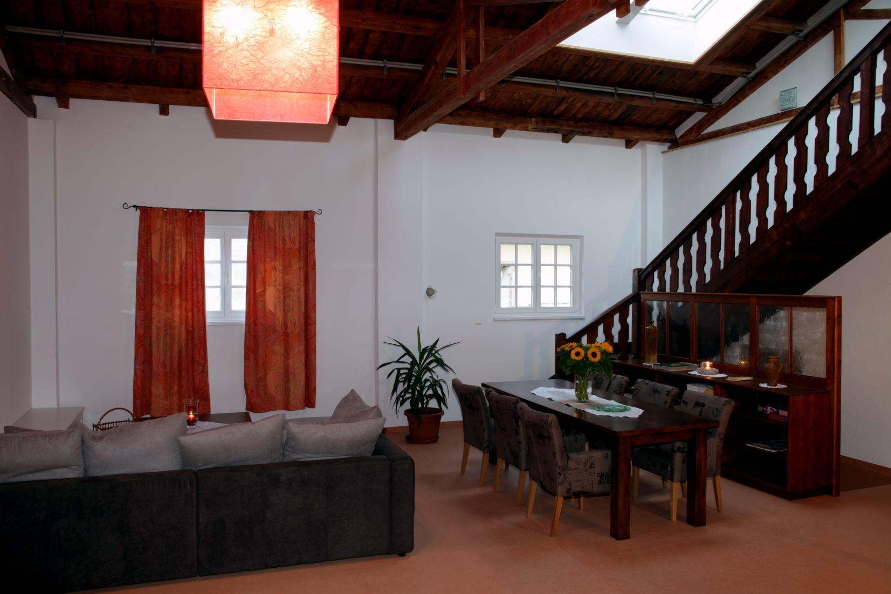 Wohn- und Essbereich in einem Loft mit Holzbalkendecke, rotem Lampenschirm, grauem Sofa, Esstisch mit Stühlen und Treppe.