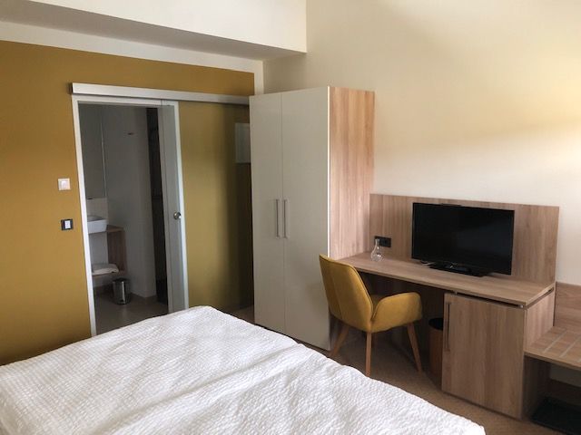 Ein modernes Gästezimmer mit Bett, Schreibtisch, Stuhl, Fernseher und Schrank. Im Hintergrund ist ein Badezimmer zu sehen.