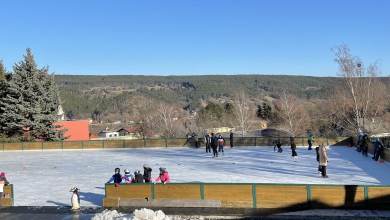 Eislaufplatz mit Menschen, umgeben von Bäumen und Hügeln unter blauem Himmel.