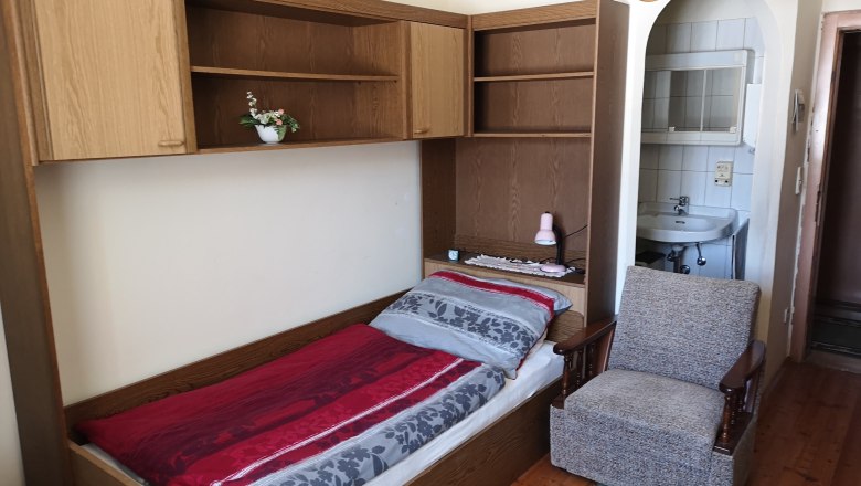 Single bed with red and gray bed linen, an armchair next to it and a washbasin in the background.