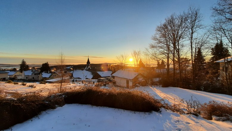 Winterlandschaft mit Sonnenaufgang über einem verschneiten Dorf und kahlen Bäumen.