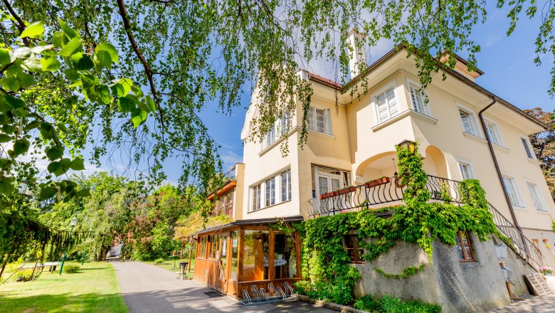 Herrenhaus Woltron, © Wiener Alpen / Christian Kremsl Ein gelbes Herrenhaus mit Efeu bewachsen, umgeben von Bäumen und einem Gartenweg.
