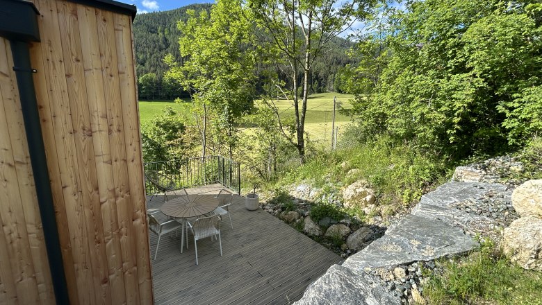 Terrasse mit Holztisch und St&uuml;hlen, umgeben von B&auml;umen und Wiese, mit Blick auf bewaldete H&uuml;gel.