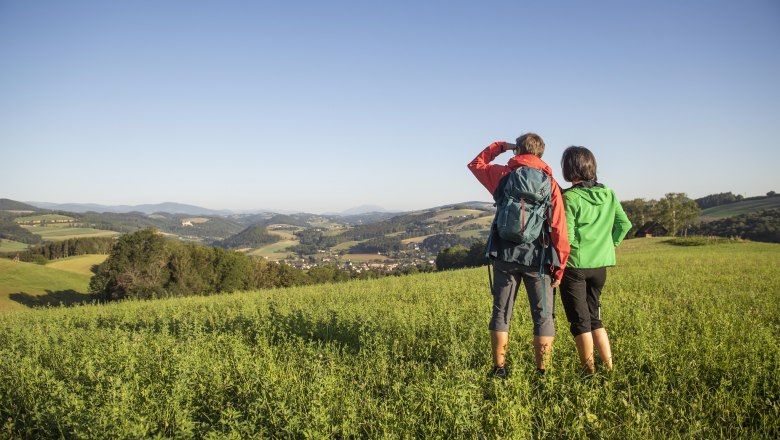 Zwei Personen stehen auf einer Wiese und blicken in die Ferne &uuml;ber eine h&uuml;gelige Landschaft.
