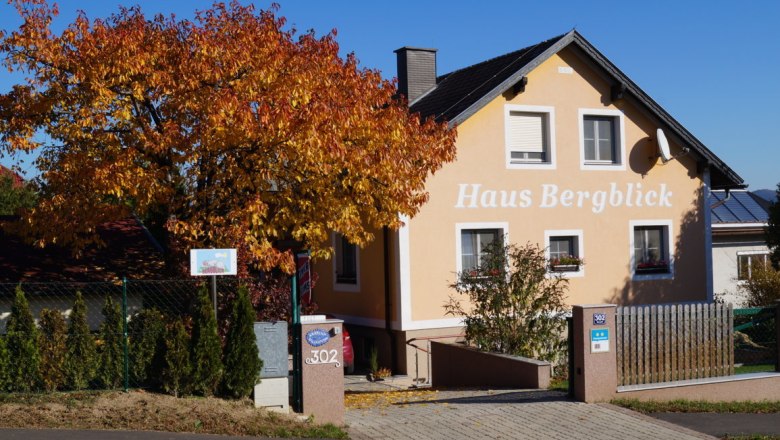 Ein gelbes Haus mit der Aufschrift 'Haus Bergblick' und einem herbstlichen Baum davor.