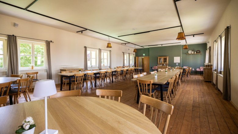 Ein großer, heller Saal mit Holzböden und Holztischen und -stühlen. Fenster an der linken Wand lassen Tageslicht herein. Der Saal ist modern gestaltet, eine Wand grün gestrichen, der Rest weiß.