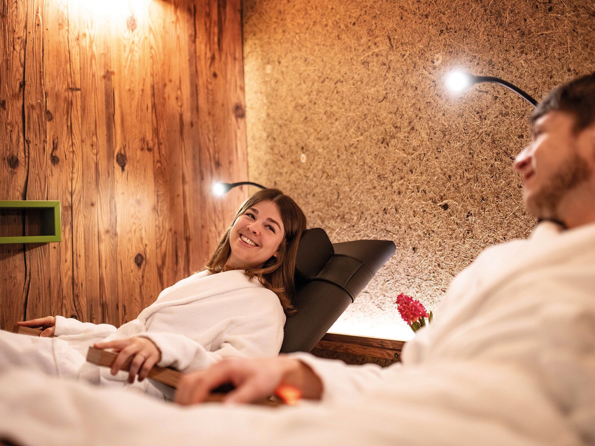 In einem einladenden Wellnessbereich entspannen zwei Gäste in flauschigen Bademänteln, umgeben von warmem Licht und einer beruhigenden Atmosphäre. Ihre Gesichter strahlen Zufriedenheit aus, während sie die Ruhe und den Komfort genießen, die dieser Ort bietet. Hier wird der Stress des Alltags vergessen und die Seele baumeln gelassen.