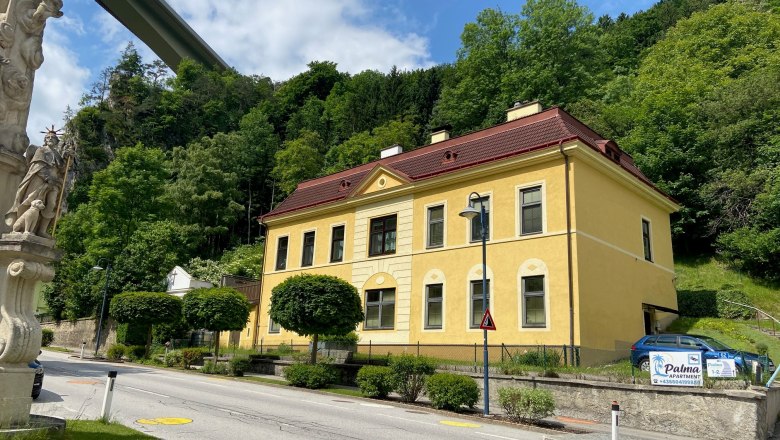Gelbes Gebäude mit rotem Dach neben einer Straße, umgeben von Bäumen und einer Statue im Vordergrund.
