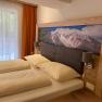 Ein gem&uuml;tliches Doppelzimmer mit einem gro&szlig;en Bett, Holzm&ouml;beln und einem Bild von Bergen &uuml;ber dem Kopfteil. Ein Fenster mit Vorh&auml;ngen ist zu sehen.