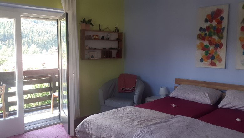 Ein gemütliches Doppelzimmer mit zwei Betten, einem Balkon mit Blick auf Bäume und bunte Kunstwerke an der Wand.