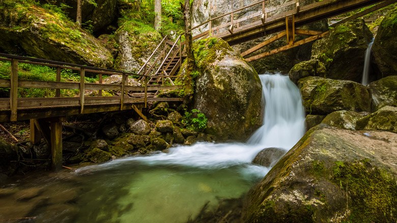 Holzbrücken und Wasserfall in einer bewaldeten Schlucht.