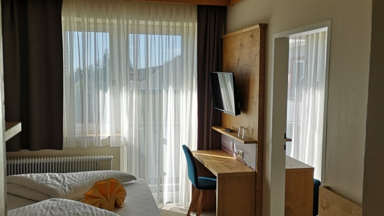 A modern double room with a bed, desk, chair and TV. Large windows with curtains let in daylight.