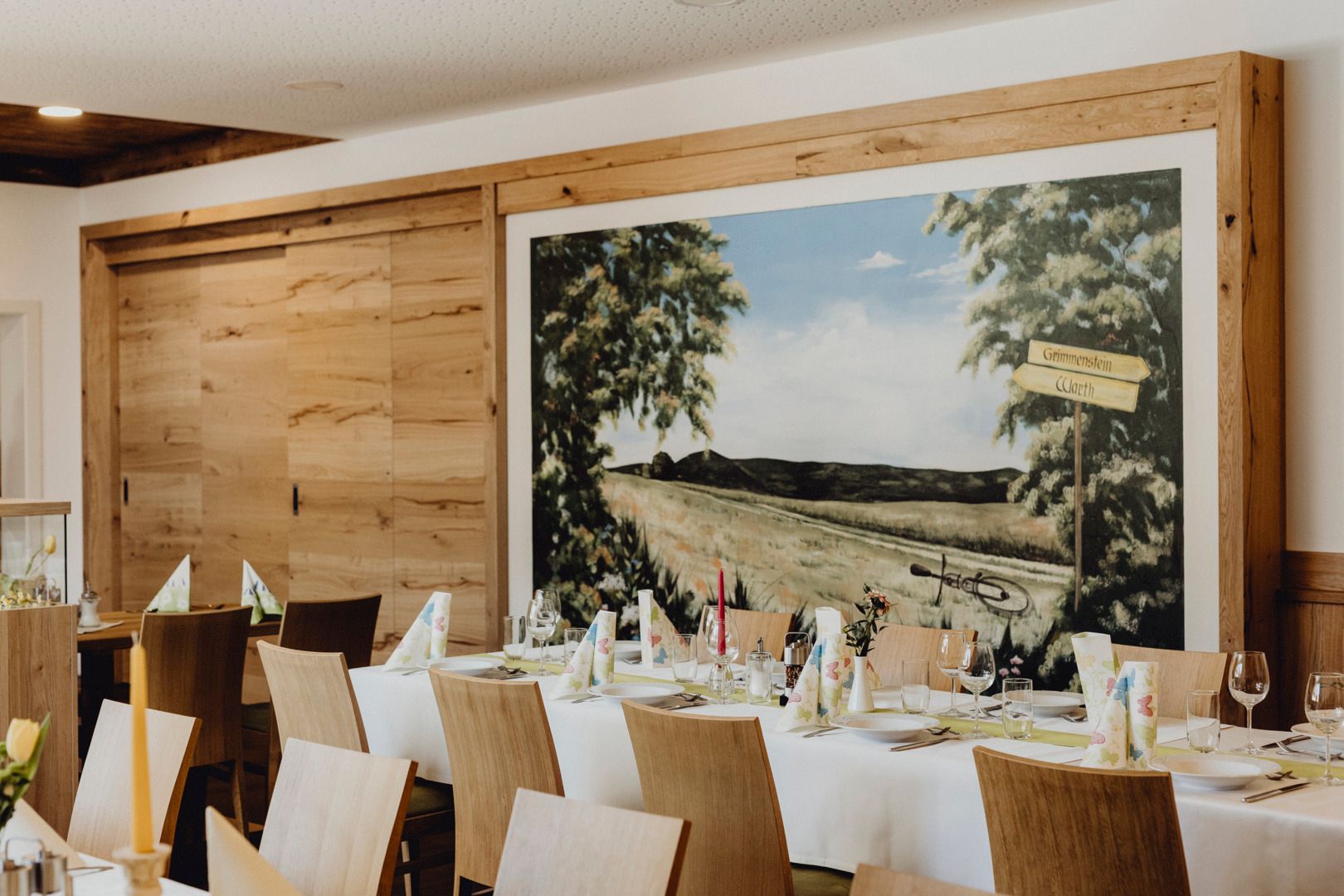 Ein heller Raum eines Gasthauses mit gedeckten Tischen, Holzstühlen und einer Holzwand mit Landschaftsbild.