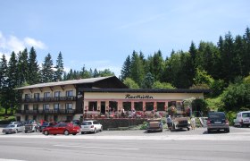 A rest house with a terrace and parked cars in front of it, surrounded by trees.