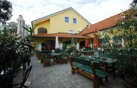Gastgarten mit grünen Tischen und Bänken sowie geschlossenen Sonnenschirmen vor einem gelben Gebäude.