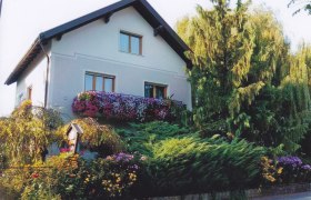 Ein zweistöckiges Haus mit Blumen auf dem Balkon und umgeben von Bäumen und Sträuchern.