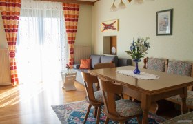 Ein gemütliches Wohnzimmer mit Holztisch, Stühlen, grauem Sofa und bunten Vorhängen. Ein Blumenstrauß steht auf dem Tisch.