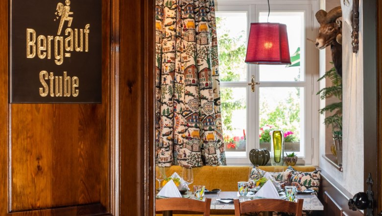 Ein gemütlicher Raum in einem Restaurant mit Holzmöbeln, einem Tisch mit gedecktem Geschirr, bunten Vorhängen und einem Geweih an der Wand.